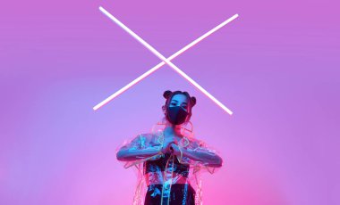 Güzel virüs maskeli Asyalı kadın portresi yüz koruması ve renkli parlak uv ışıkları etrafında yağmurluk stüdyoda poz veriyor.