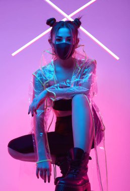 Güzel virüs maskeli Asyalı kadın portresi yüz koruması ve renkli parlak uv ışıkları etrafında yağmurluk stüdyoda poz veriyor.