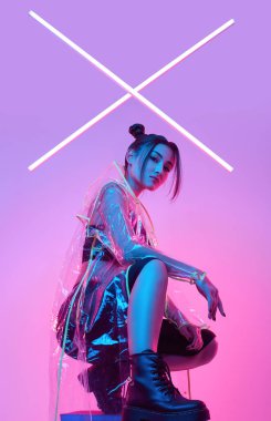 Renkli parlak neon ışıkların etrafında şık bir yağmurlukla stüdyoda poz veren zarif Asyalı bir kadının portresi.