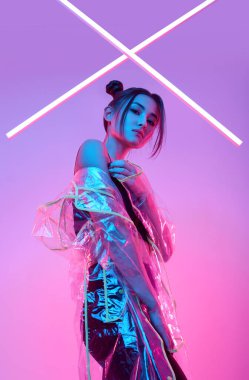 Renkli parlak neon ışıkların etrafında şık bir yağmurlukla stüdyoda poz veren zarif Asyalı bir kadının portresi.