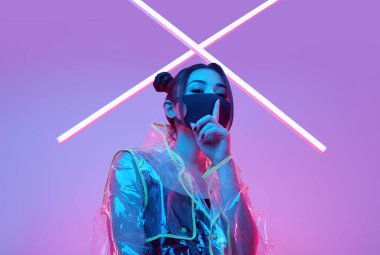 Güzel virüs maskeli Asyalı kadın portresi yüz koruması ve renkli parlak uv ışıkları etrafında yağmurluk stüdyoda poz veriyor.