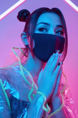 Güzel virüs maskeli Asyalı kadın portresi yüz koruması ve renkli parlak uv ışıkları etrafında yağmurluk stüdyoda poz veriyor.