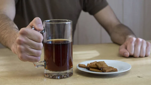 Bir adam masaya oturur ve elinde bir kupa koyu bira tutar. Masanın üzerinde bir kupa ve ekmek kırıntılarıyla dolu bir tabak var..