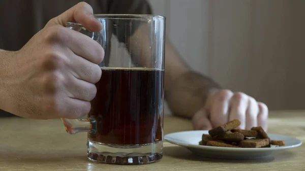 Bir adam masaya oturur ve elinde bir kupa koyu bira tutar. Masanın üzerinde bir kupa ve ekmek kırıntılarıyla dolu bir tabak var..