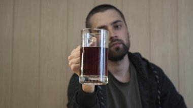 Genç, kısa saçlı, sakallı esmer bir adam cam bardaktan koyu bira içer..