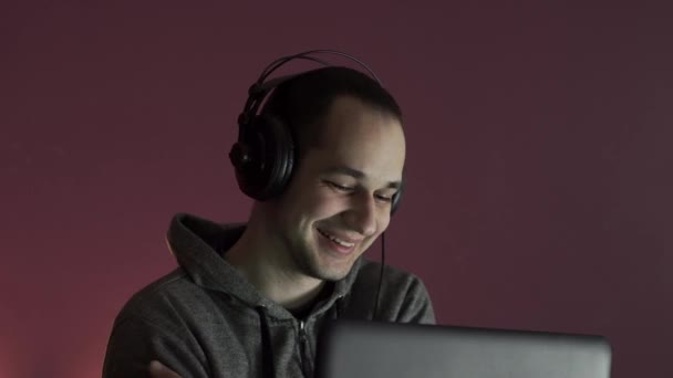 若い男がヘッドフォンでコンピュータの近くの夜に座って 友人とオンラインで話している間に笑います ホームコロナウイルスの概念を滞在 — ストック動画