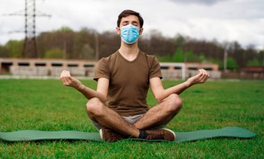Tıbbi maskeli yakışıklı bir adam stadyumun çimlerinde meditasyon yapıyor. Coronavirüs salgını konsepti.