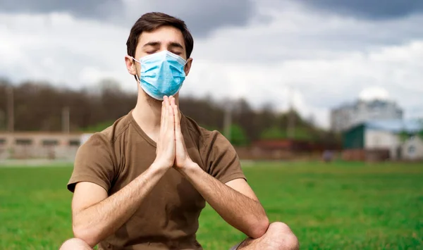 A handsome guy in a medical mask makes meditation on the grass of a stadium. Coronavirus epidemic concept.