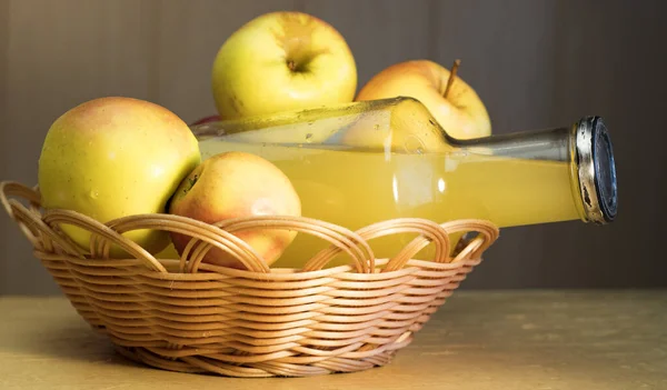 A large bottle of apple juice and fresh apples in a basket on the table.