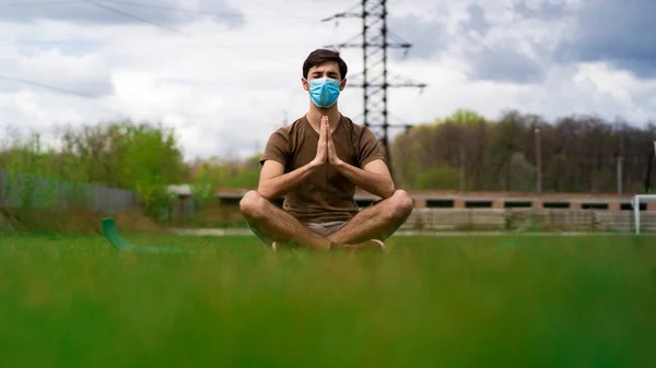 A handsome guy in a medical mask makes meditation on the grass of a stadium. Coronavirus epidemic concept.