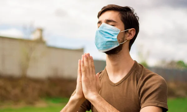 A handsome guy in a medical mask makes meditation on the green grass of a stadium. Coronavirus epidemic concept.