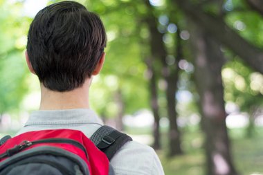 Genç bir erkek turist gri-kırmızı bir sırt çantasıyla parkta yürüyor. Arkadan bak. Kopya alanı ile doğa yolculuğu kavramı.