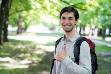 Gri tişörtlü, sırt çantalı, parkta durup gülümseyen güzel bir beyaz adam. Kopya uzayı ile seyahat kavramı. Yakışıklı adam başparmağını kaldırıyor. Turist programları....