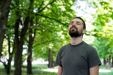 Güzel, genç bir esmer parkta dikiliyor, başını yukarı kaldırıyor ve gözlerini kapıyor ve gülümseyerek hayatın tadını çıkarıyor..