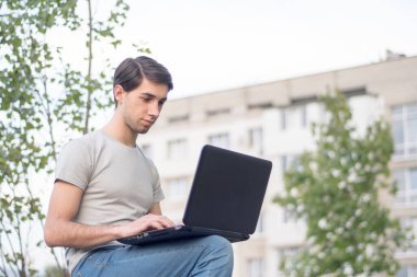 Parkta oturan ve dizüstü bilgisayarda yazan beyaz bir genç. Uzak çalışma konsepti