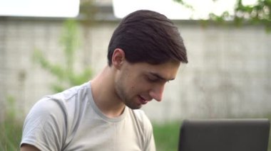 Genç beyaz bir adam evin yakınındaki çimlerin üzerinde oturuyor ve bir dizüstü bilgisayarla yazı yazıyor. Uzak çalışma konsepti, kapat.