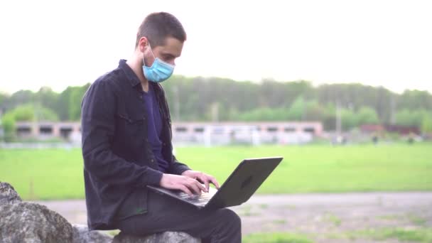医療用のマスクをした若い男がスタジアムの近くの石の上に座っていて すぐにノートパソコンを入力しています リモートワークの概念 — ストック動画