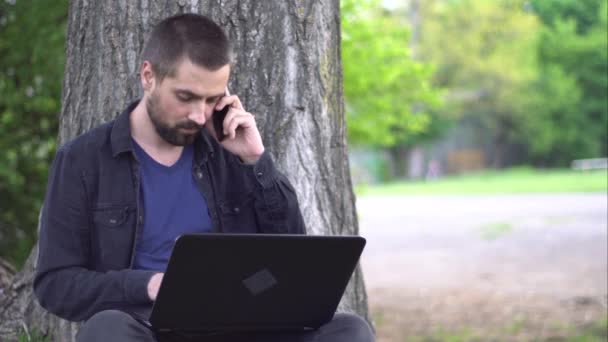 髭を生やした若い白人男性が公園の木の下に座り ノートパソコンをタイプして電話で話している リモートワークの概念 — ストック動画