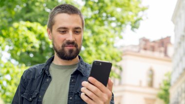 Sakallı, genç, beyaz bir adam uzanmış elinde bir telefon tutuyor ve selfie çekiyor. Blogcu görüntülü arama ile iletişim kuruyor. Adam caddede gülümsüyor.