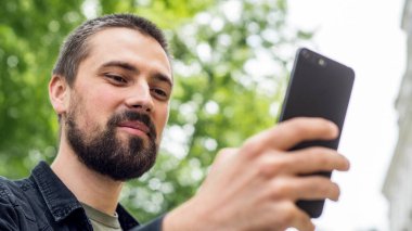 Sakallı, genç, beyaz bir adam uzanmış elinde bir telefon tutuyor ve selfie çekiyor. Blogcu görüntülü arama ile iletişim kuruyor. Adam caddede gülümsüyor. Portreyi kapat.
