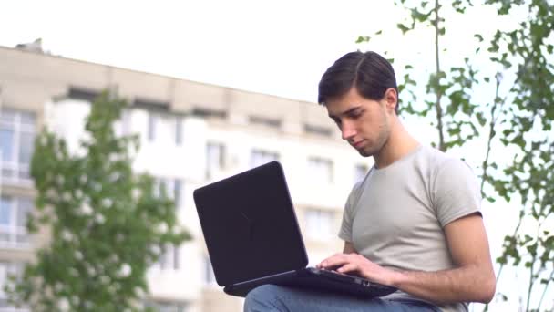 若い白人男性が家の近くの芝生の上に座ってノートパソコンをタイプしています リモートワークの概念 — ストック動画