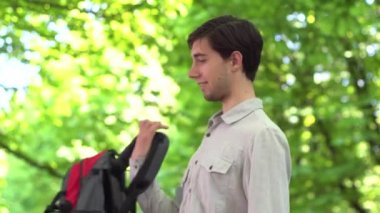 Parkta genç beyaz bir adam sırtına bir turist çantası koyar ve gülümser..