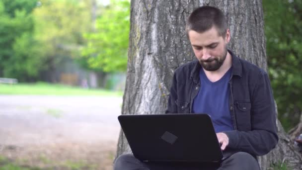 髭を生やした若い白人男性が公園の木の下に座ってノートパソコンをタイプしている リモートワークの概念 — ストック動画