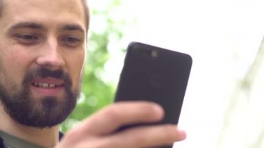 Sakallı, genç, beyaz bir adam elinde bir telefon tutuyor ve konuşuyor. Blogcu görüntülü arama ile iletişim kuruyor. Adam caddede gülümsüyor. Kapat..