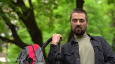 Sakallı yakışıklı beyaz bir adam omzuna bir sırt çantası atıyor. Parkta sırt çantalı bir yürüyüşçü.