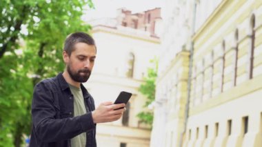 Sakallı, beyaz bir adam elinde telefon olan bir şehir caddesinde duruyor. Önce ciddi ve odaklanmış bir şekilde küfrediyor, sonra gülümsüyor ve selfie çekiyor..