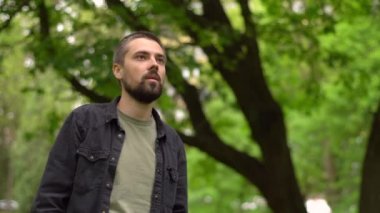 Genç sakallı beyaz bir adam parkta durur ve yüzünü peçeteyle kapatarak hapşırır. Bahar alerjisi konsepti.