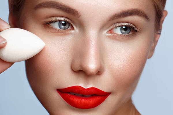 Beautiful girl with red lips and classic makeup with foundation and sponge in hand. Beauty face.