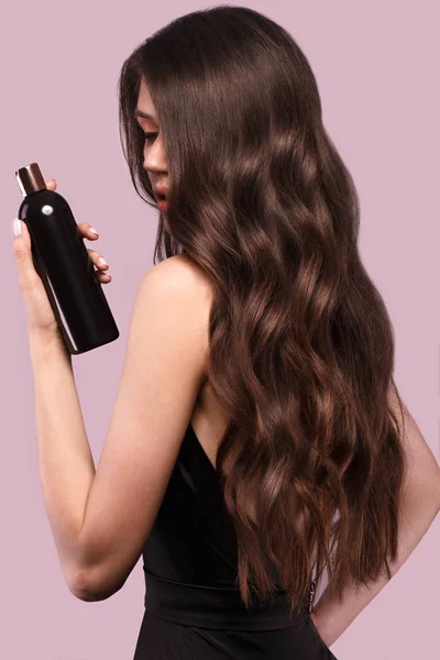 Hermosa morena con rizos y maquillaje clásico en un vestido negro con una botella de champú en las manos. Cara de belleza . —  Fotos de Stock