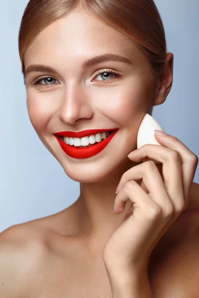 Piękna dziewczyna z czerwonymi ustami i klasycznym makijażem z gąbką fundacji w dłoni. Piękna twarz. — Zdjęcie stockowe
