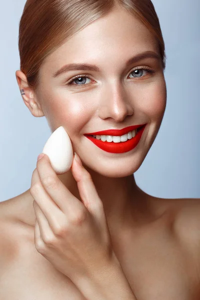 Piękna dziewczyna z czerwonymi ustami i klasycznym makijażem z gąbką fundacji w dłoni. Piękna twarz. — Zdjęcie stockowe