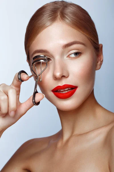 Vacker tjej med röda läppar och klassisk makeup med locktång i handen. Skönhetsansikte. — Stockfoto
