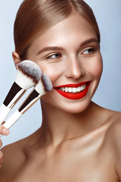 Mooi meisje met rode lippen en klassieke make-up met cosmetische borstels in de hand. Schoonheidsgezicht. — Stockfoto