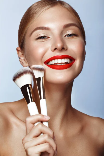Piękna dziewczyna z czerwonymi ustami i klasycznym makijażem ze szczotkami kosmetycznymi w ręku. Piękna twarz. — Zdjęcie stockowe