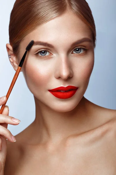 Vacker tjej med röda läppar och klassisk makeup med kosmetisk brynborste i handen. Skönhetsansikte. — Stockfoto