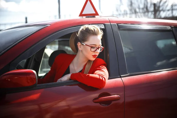 Νεαρή όμορφη ευτυχισμένη γυναίκα στο κόκκινο κάθεται στο τιμόνι νέο εκπαιδευτικό αυτοκίνητο. — Φωτογραφία Αρχείου