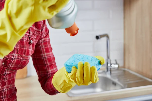 Women's hands in yellow protective glove with bottle of dishwashing liquid and sponge on kitchen. Cleaning concept. — Stock fotografie