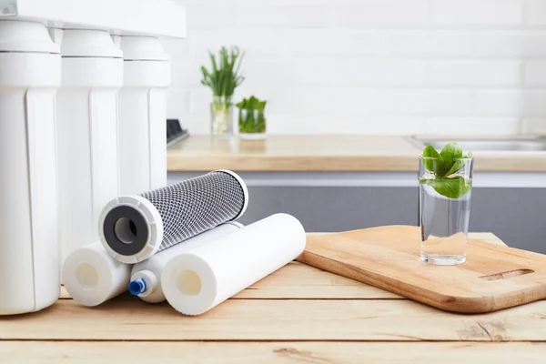 A glass of clean water with osmosis filter and cartridges on wooden table in a kitchen. Concept filtration system.