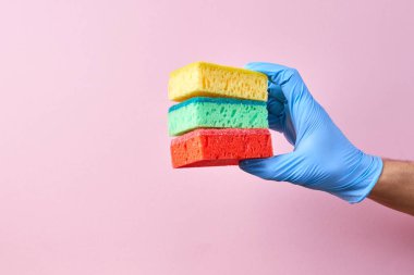 Hand in blue rubber protective glove holding different: yellow, green and red sponge on pink background