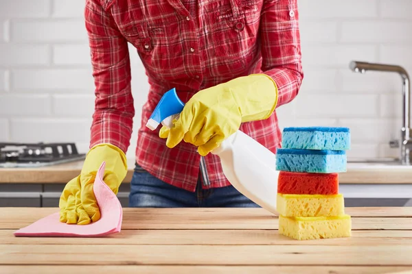 Wanita dalam sarung tangan karet kuning membersihkan meja kayu dengan semprotan, spons dan kain, latar belakang dapur — Stok Foto