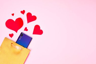 Valentines day flat lay copy space. Golden paper gift bag, smartphone And red hearts on pink paper background.
