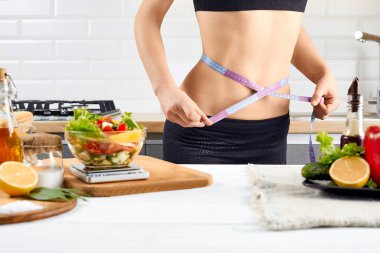 Diyet, sağlıklı beslenme, yemek ve kilo kaybı konsepti. Mutfağın içinde domates, biber ve salata yanında belini ölçen genç bir kadın var..