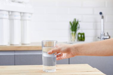 Çocuk el ahşap bir masadan bir bardak arıtılmış su alır. Arka planda Osmosis filtresi ve musluk. Su tedavisi kavramı