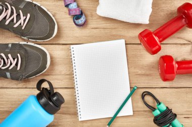 Fitness and healthy active lifestyle background concept. Athlete's equipment: dumbbell, water bottles, notepad, jump rope, sneakers, on natural wooden background. Flat lay. Space for text.