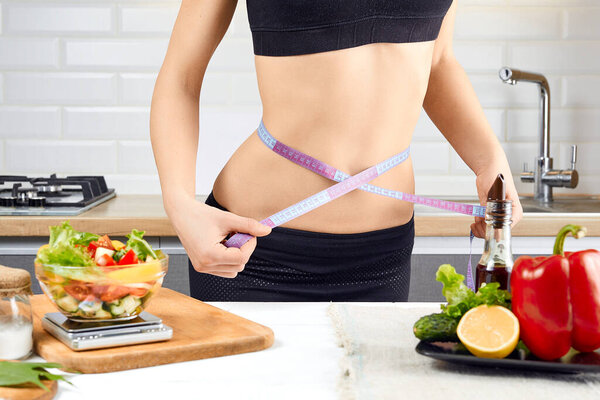 Young sports girl with naked torso measuring her waist with measuring tape in the kitchen. Ripe tomatoes, peppers and salad on the table. diet and weigh loss concept