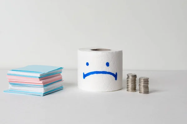 Toilet paper roll with sad smile and coins on a white background. Covid-19 and coronavirus panic concept